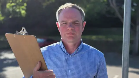 Paul Moseley/BBC Andrew Sykes, a man who is standing outside holding a wooden clipboard. He is looking directly at the camera and is wearing a blue checked shirt.