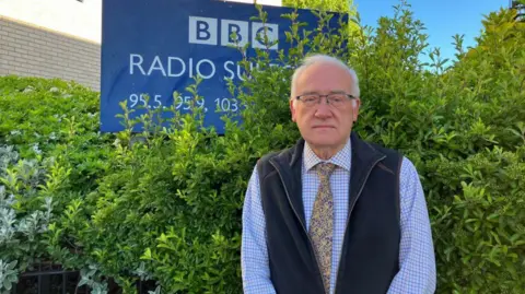 Vikki Irwin/BBC Christopher Hudson is standing in front of the BBC Radio Suffolk building. There is a hedge to the right in the background and to the left there is a BBC Radio Suffolk sign. Christopher is wearing a patterned blue shirt, yellow tie and dark blue fleece gilet. 