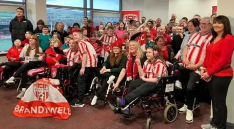 A number of people are seen in Derry City's stadium hospitality suite.
They are predominantly wearing red and white Derry City jerseys.
A fla with Derry City's club crest can be seen near the front of the group.