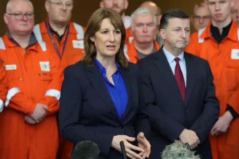 PA Media The Chancellor Rachel Reeves and Scottish Secretary Douglas Alexander at the Grangemouth plant. Ms Reeves has short brown hair and a blue jacket and top. Mr Alexander is wearing a blue suit and maroon tie. Workers from the factory in orange clothing stand behind them