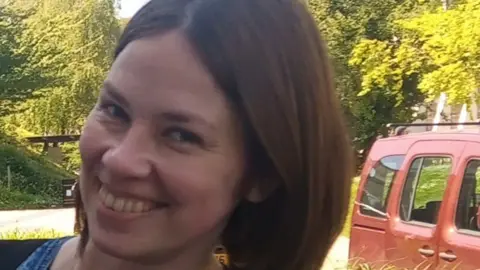 South Wales Police Tetiana Martynova, 40, from Kharkiv, Ukraine, smiles at the camera. She has a short brown bob and brown eyes. Green trees can be seen behind her, as well as part of a red car. 