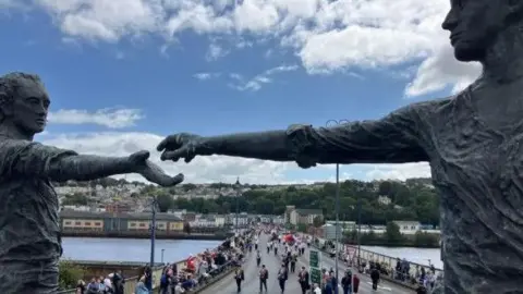 A statue of two people reaching out for each others hands. The Craigavon Bridge can be seen with members of the Apprentice Boys of Derry marching across the bridge with spectators either side. 