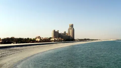 Ras Al Khaimah beach, which is sandy and has a clear blue sea. There are a few tall buildings scattered along the coast.