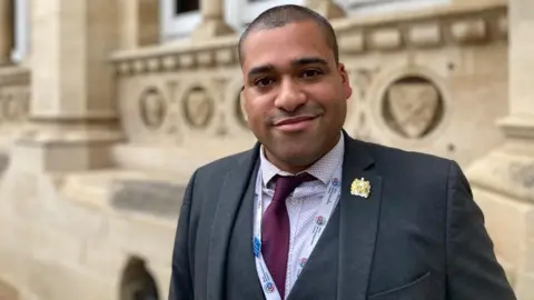 Laura Coffey/BBC James Hill, standing outside, wearing a burgundy tie, lilac patterned shirt, and dark grey suit with a waistcoat. He has a badge on his lapel and a lanyard round his neck. He has very short dark hair and is smiling and looking straight at the camera. 