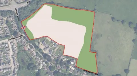 A map with a section highlighted in green and white, with a red line drawn around it. There is a built up area to the left of it and open countryside with fields and trees to the right.