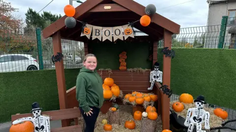 Katie Underwood A young girl in a green jumper stood in front of a pumpkin display with a Halloween sign reading 'BOO'.