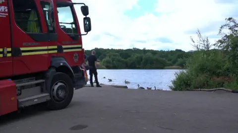 A mainly red fire engine is in the foreground on the left of the photo. A man wearing black with his back to the camera is standing between the fire engine and the water in the background. Trees are in the distance.