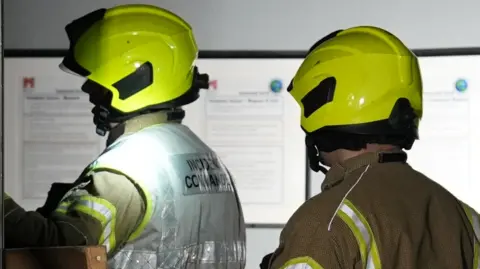 Two firefighters are moving through a dimly lit building. They are wearing bright yellow safety helmets.