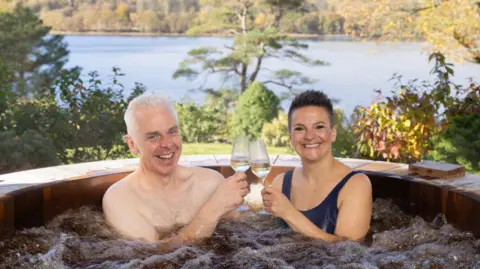 Omaze A man and a woman in a hot tub clinking champagne glasses and smiling broadly with a backdrop of Lake Coniston in the Lake District on a sunny day.