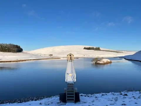 Terry Callow North Esk Reservoir in the Pentland Hills. A blue reservoir surrounded by low snowy hills and a blue sky. A bridge stretches out to a little house at the other end of the reservoir.