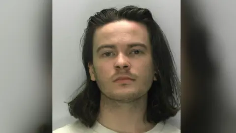 A mugshot of Samuel Rust, he is young and has shoulder length black hair, a nose piercing and an ear piercing on his right ear.