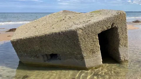 The pillbox is a block-shaped concrete structure. It is lying, partly buried in sand.