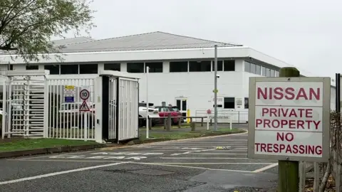 The security gates at the front of the Nissan building, with white gates open for vehicles and a carousel gate for pedestrians. There is a sign with red writing that says "Nissan / Private Property / No Trespassing". The building is about three storeys tall with a pointy silver roof.