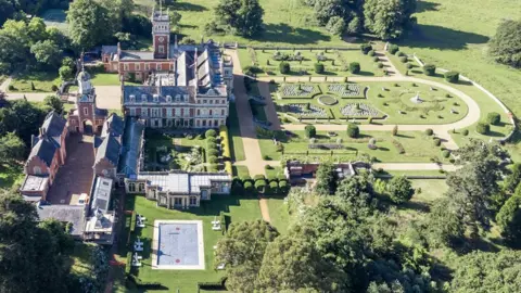 Historic England Archive An aerial shot of Somerleyton Hall and gardens. The house is on the left and is a u-shaped, three storey mansion. Running down from its lower wing is a walled garden with a long run of glasshouses on the left. To the right can be seen formal parterre-style gardens with yew hedges.
