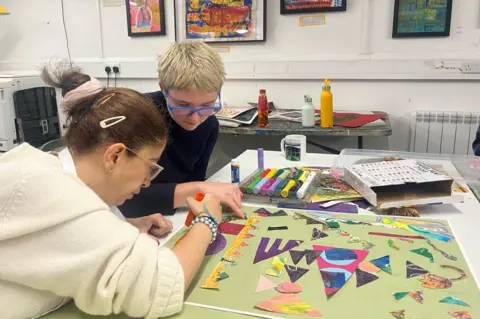 BBC / Rosa Rahimi Two women sat at a table in an art studio, working on a collage on a light green piece of paper. They are sticking multi-coloured pieces of paper onto the background.