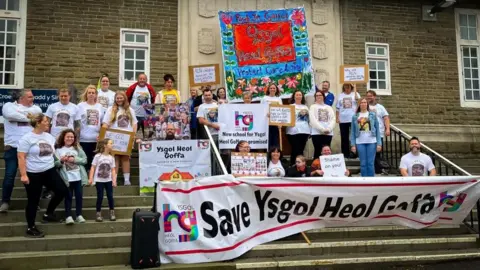 About 40 campaigners wearing white shirt hold up signs as they stand on the steps of Carmarthen council. A main banner says Save ysgol heol goffa 