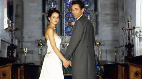 A bride and groom stand hand in hand inside a church, a large stained-glass window behind them. The bride has dark hair piled up and is wearing a strapless white dress, standing side on, looking to camera. The groom has his back to the camera and is turning around and smiling. He is wearing grey tails and trousers. Wooden pews and brass church ornaments can be seen around them.