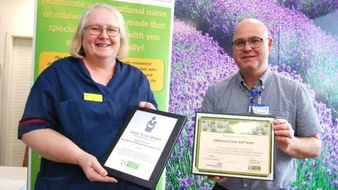 Kettering General Hospital Jo Snow with medium length blond hair and glasses, wearing a dark blue uniform, holds a certificate in a black frame. Robin Binks, with very short hair and glasses, wearing a blue and white shirt and a lanyard, holds another certificate in a black fraime. They are both standing in front of a big picture showing purple flowers in a field.