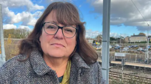 Nicola Haseler/BBC Sarah Conboy is on the left of the image stood in front of a train station. She had shoulder length brown hair and was wearing glasses, a green jumper, and tweed coat. 