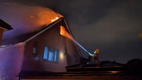 Dave Marubbi A house, at night, with its roof on fire and a firefighter spraying water on the flames with a hose jet.