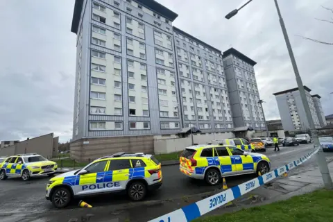 BBC A block of flats clad in blue and white. Police cars line the residential streets and there is police tape blocking off the area, tied to a lamppost. 