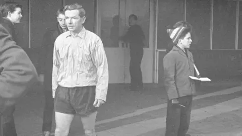 Getty Images A black-and-white image of a man in a long-sleeved shirt, shorts and football socks walks along a paved area in front of a glass-fronted building. A child with an autograph book stands several paces away, looking away.