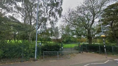 Google One of the entrances to Saltwell Park in Gateshead. A zebra crossing (just in shot on the right hand side) leads to a green pair of gates set into a green metal spiked fence. Behind it is an expanse of green grass and several trees. There is a buckled, grey metal street barrier just outside the gates and a wooden sign to the left. It is an overcast day but the trees are in full leaf.