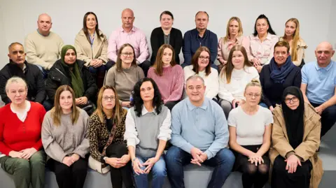 Twenty-three people, men and women, sit in three rows and look solemnly at the camera. They are in a white room sitting on a grey platform. Most of the people at the front have their hands clasped over their knees.