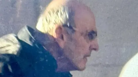 A blurred shot of a man with white hair and glasses seen from the side as he arrives at court. He is wearing a thick black coat with hood.
