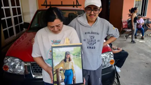 Relatives of América Sánchez hold up a framed photograph of the young cadet in her hometown of Xalapa. in the photo, América Sánchez, is posing in a yellow blouse and jeans in front of a lake. Her relatives are crying.