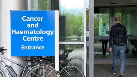 Dan Freeman walking into a door at the Churchill Hospital which has a blue and white sign next to it saying 'Cancer and Haematology Centre'.