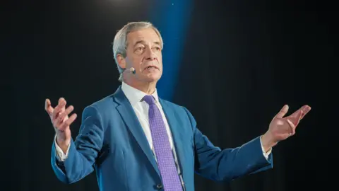 Reform UK leader Nigel Farage, a middle aged man with grey hair, a white shirt, blue suit, and purple tie, delivers a speech on a dark stage with a light shining down on him.