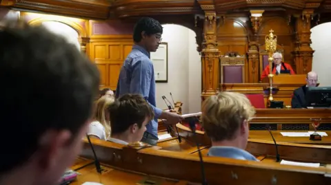States of Jersey A young man in a blue shirt with dark hair and glasses stands in the debating chamber of the States of Jersey, addressing the wood-panelled room. A gold mace is seen on the right of the picture.