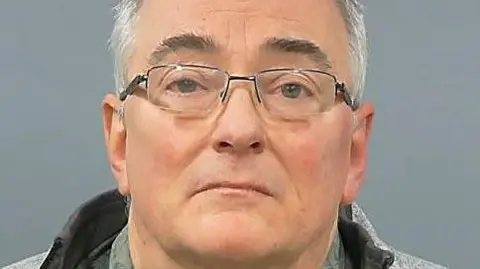 Gerard Hutchings faces the camera in a police custody photo. He has short, grey hair and wears rectangular spectacles and a grey fleece.