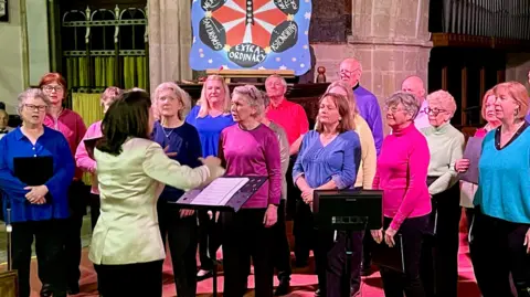 Clash of the Choirs Mainly adult female choir singers, with three men, face the choir master, who has her back to the camera. The singers range in age and are all wearing brightly coloured tops, ranging from bright blue, to pink, purple and green. In the backgrund, the clap-o-meter is visible, with some of its words showing: astonishing, extraordinary, and sparkling.