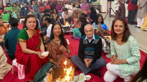 Bharat Hindu Samaj Two woman dressed in sarees and a man in a jumper and trousers and another woman in a green shirt and white trousers sitting in a semi-circle praying in front a fire - inside a community hall with more worshippers in the background.