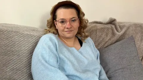 Sarah Wilson A woman with blonde curly hair looks at the camera while sitting on a grey sofa. She wears a light blue jumper.