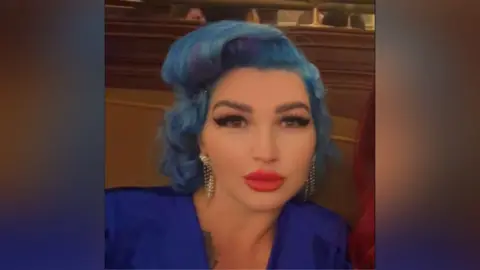 Facebook Tania Williams is inside looking to the camera. She has wavy blue hair and make-up on, including bright red lipstick and black mascara. She is wearing a dark blue dress.