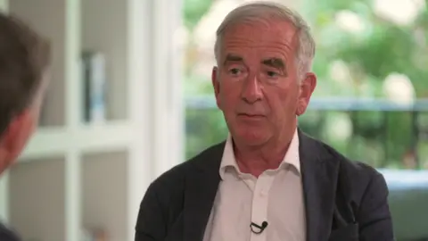 BBC Robert Harris zit in een heldere kamer met een balkon en bomen zichtbaar op de achtergrond. Hij draagt een donkere blazer en een wit shirt.