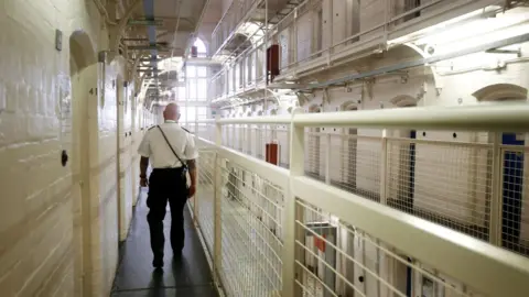 PA Media A prison officer walks along a landing at Barlinnie Prison. You can see the back of him. There is white brickwork, railings and cell doors