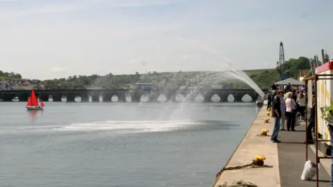 Torridge District Council A fountain going into the river. There is a small sailing boat with red sails. There is a bridge going across the river. The sky is light blue. The water is a darker blue and the fountain water is clear. There are people walking along on the right.