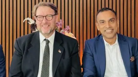 Two men are sitting next to each other shoulder to shoulder, smiling at the camera.