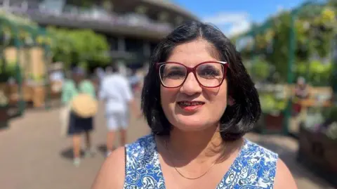 Kam Soomal is wearing a blue dress and a necklace. She has short black hair and glasses. There are people walking around in the background.