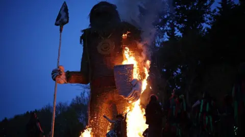 Getty Images A huge wicker figure burns at night. It is carrying a large spear. 