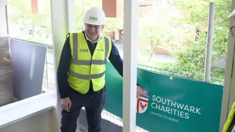 Sir Ed Davey in a hard hat and a yellow high vis tabard over a dark clothing is on a builing site, pointing at a sign that says "Southwark charities".