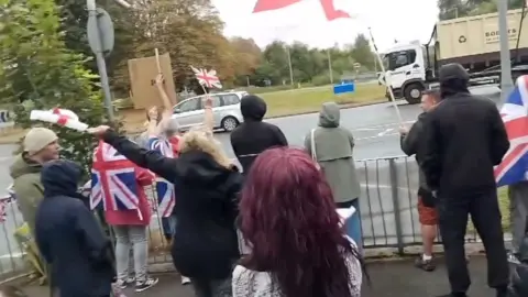 Protests in Warrington, people wave Union flags. About 10 people are standing, their backs to the camera. Some are waving England or Union flags. Traffic is driving past.