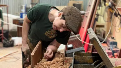 Contributed A woman working in a boat yard workshop. She is working on a piece of wood and is leaning over a workstation. She wears a flat cap, green T-shirt and brown trousers. 