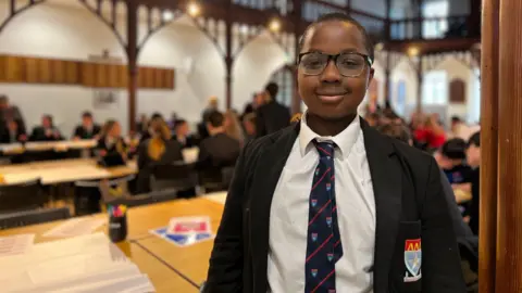 Alex smiles at the camera. He is wearing a black blazer with the school logo on his chest and a dark blue tie with red diagonal stripes alongside the school logo. He has a white shirt and black frame glasses.