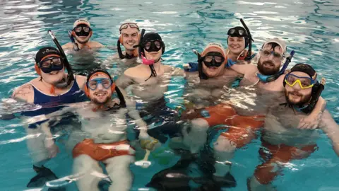 A mixed gender group of people pose with their arms around each other in a swimming pool. Everybody wears black snorkels, swimming caps and masks. The three people at the front can be seen wearing bright orange swimming trunks and flippers.