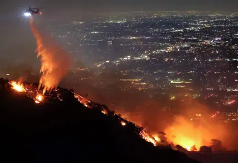 Mario Tama/Getty Images A firefighting helicopter drops water as the Sunset Fire burns in the Hollywood Hills at night on Wednesday, with a view of the Los Angeles skyline in the background.